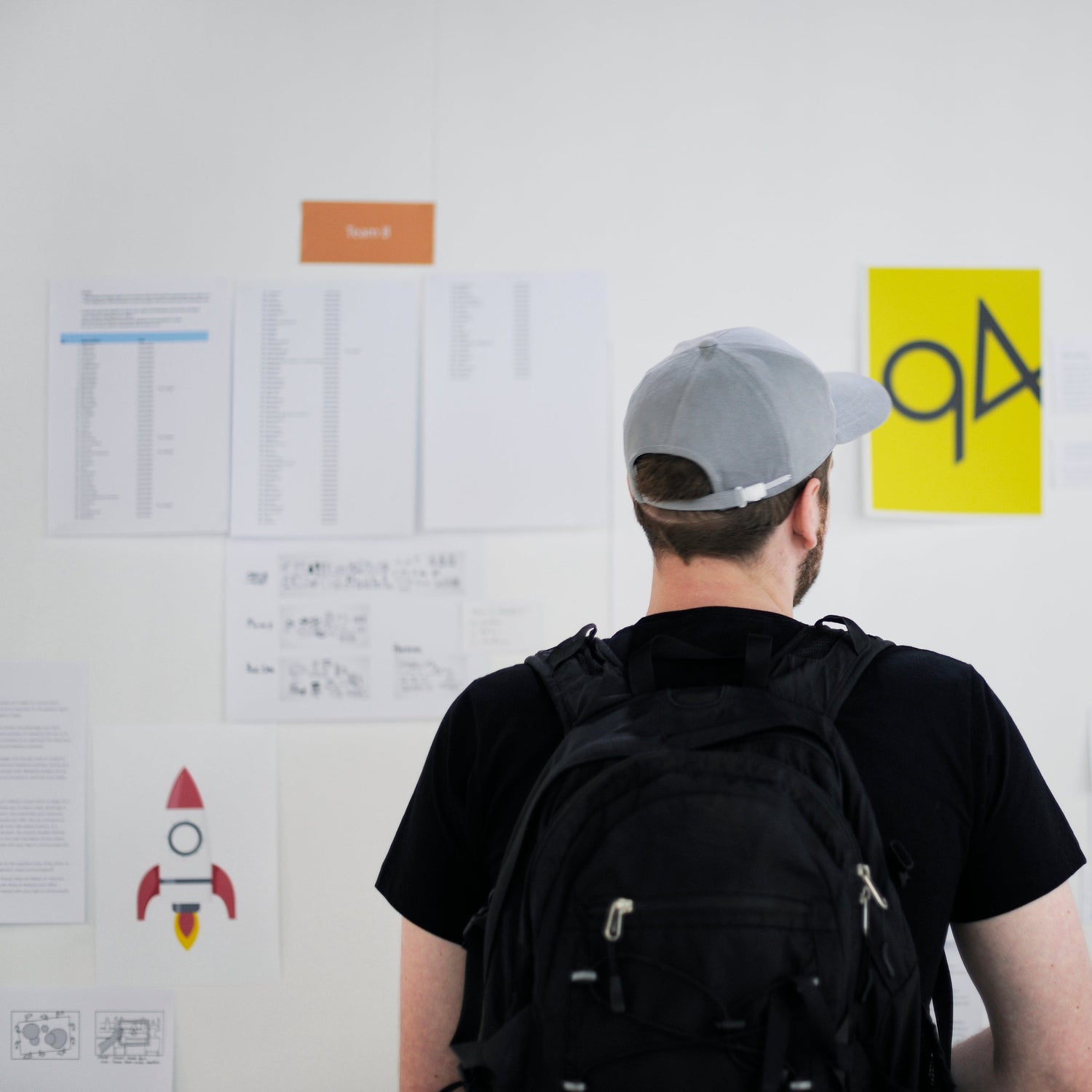A student wearing a backpack and a gray cap looks at a wall filled with design sketches, brainstorming creative ideas for modern furniture concepts.